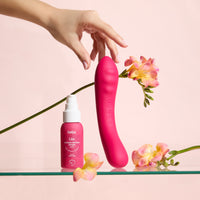 Vibratore rosso Double Trouble e lubrificante Liio su superficie trasparente, con fiori e mano che tocca il prodotto, sfondo rosa tenue