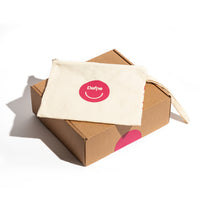 Pochette Dafne in cotone naturale con logo rosso, appoggiata su scatola in cartone, su sfondo bianco